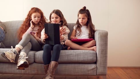 Foto: Colourbox Tre barn sitter på en sofa og bruker digitale enheter. Det ene barnet til venstre holder en smarttelefon, barnet i midten leser på en nettbrett, og barnet til høyre holder et annet nettbrett. De sitter komfortabelt i et hjemmemiljø med en nøytral farget bakgrunn.